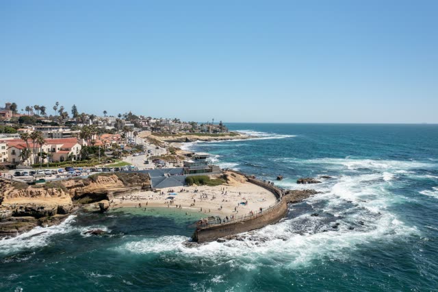 Aerial view of the cliffs, the surf and palm trees at La Jolla Cove, with a crowd gathered.