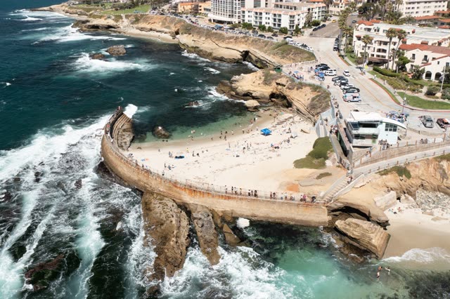 Aerial view of the cliffs, the surf and boats at La Jolla Cove, with a crowd gathered.