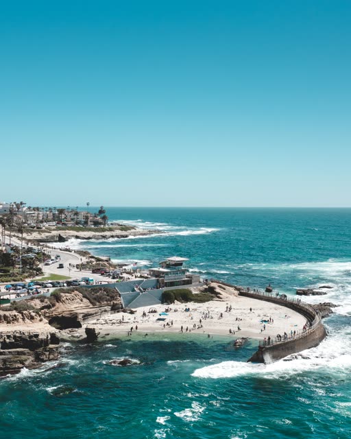 Aerial view of the cliffs, the surf and palm trees at La Jolla Cove, with a crowd gathered.