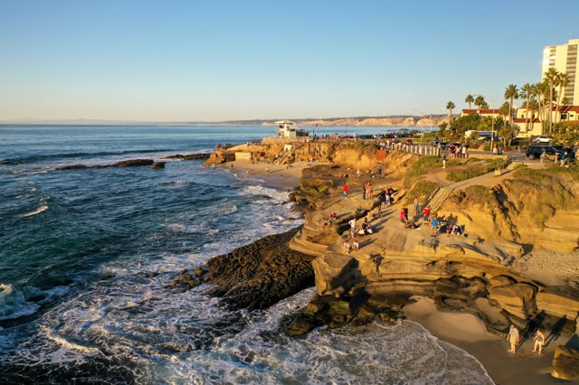 Low aerial view of the cliffs, the surf and palm trees at La Jolla Cove, with people walking.